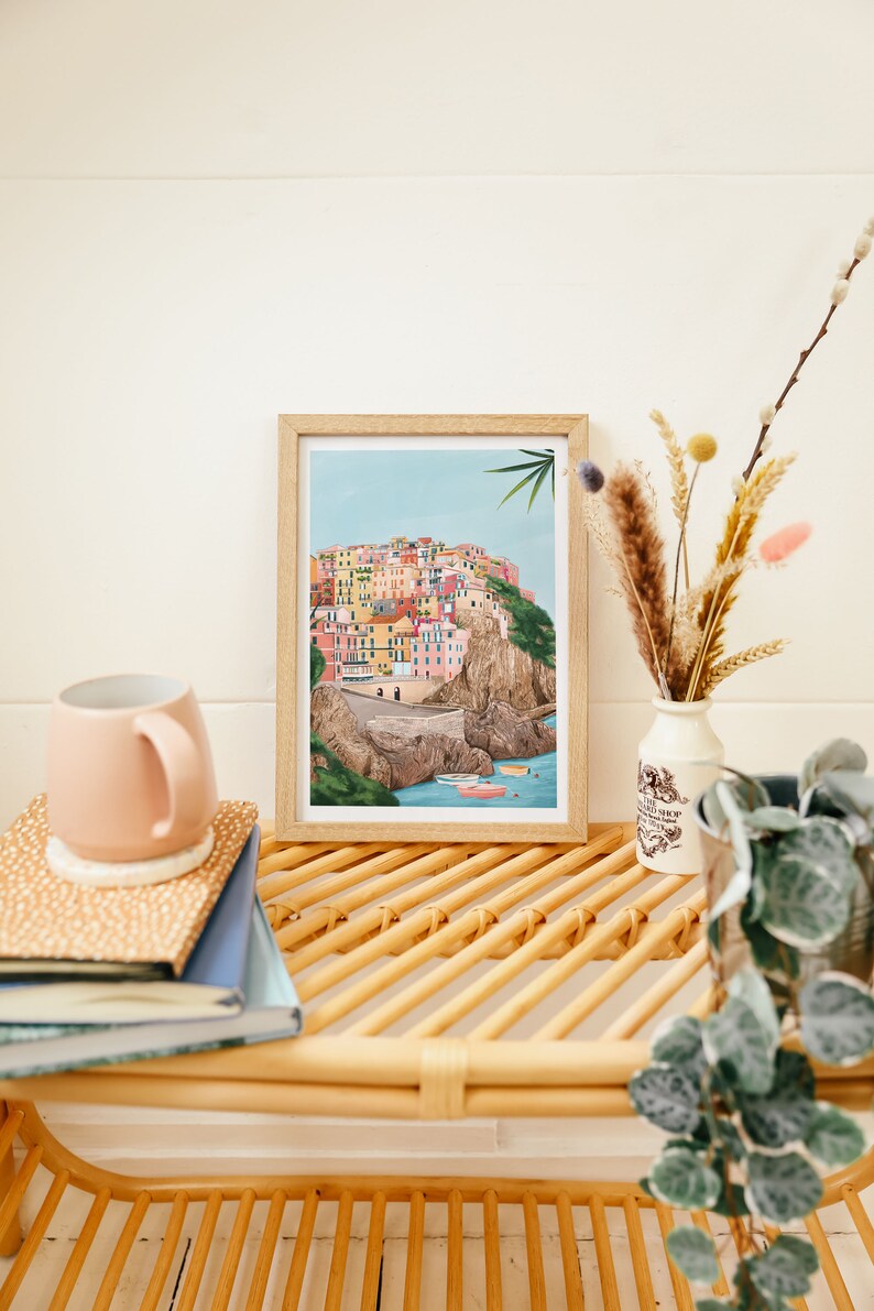 May include: A framed art print depicting a colorful coastal village scene. The artwork features buildings in various colors, set against a blue sky and sea. A pink mug, books, and decorative vase with dried flowers are also visible.