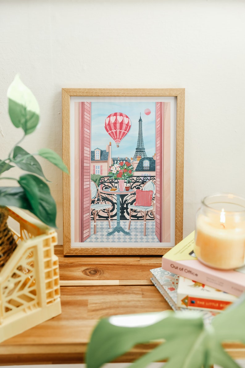 May include: A framed illustration of a Parisian balcony scene with a hot air balloon, Eiffel Tower, and a table with two chairs. The balloon is pink and white with hearts, and the Eiffel Tower is in the background. The balcony is decorated with pink flowers.