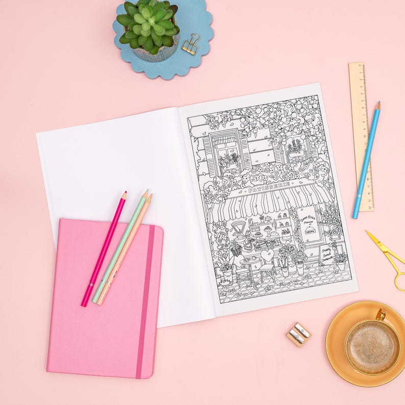 May include: An open coloring book with a detailed black and white illustration of a French patisserie. A pink notebook, colored pencils, and a cup of coffee are arranged on a pink surface. A small succulent plant sits on a blue scalloped tray.