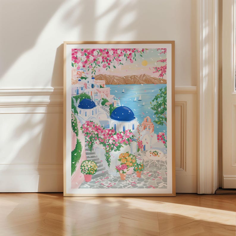 以下が含まれることがあります： 白い建物、青いドーム、ピンクの花が描かれた、海岸沿いの村の風景の額装されたアートプリント。このアートワークは、晴れた空、ヨットのある穏やかな海、石畳の小道が特徴です。プリントは明るい木製のフレームに入っています。