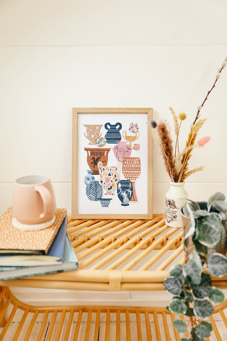 May include: A framed print of a colorful illustration featuring various stylized ceramic vases and urns. The print is displayed on a light wood shelf with a mug, books, and dried flowers.