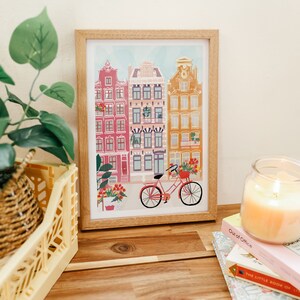 Puede incluir: Una ilustraci&oacute;n enmarcada de una escena de calle con edificios coloridos y una bicicleta con flores en la cesta. Los edificios tienen fachadas rojas, naranjas y amarillas con molduras blancas. La bicicleta es roja con una cesta blanca y ruedas blancas.