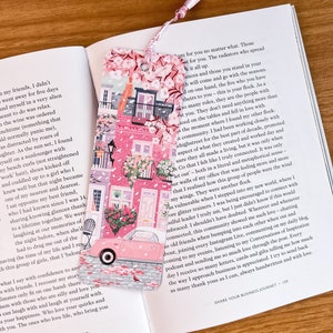 Pode incluir: Marcador de p&aacute;gina rosa e branco com um carro rosa e um design floral. O marcador mostra um carro rosa dirigindo por uma rua com edif&iacute;cios rosa e flores. O texto no marcador diz "Fa&ccedil;a o que ama, ame o que faz".