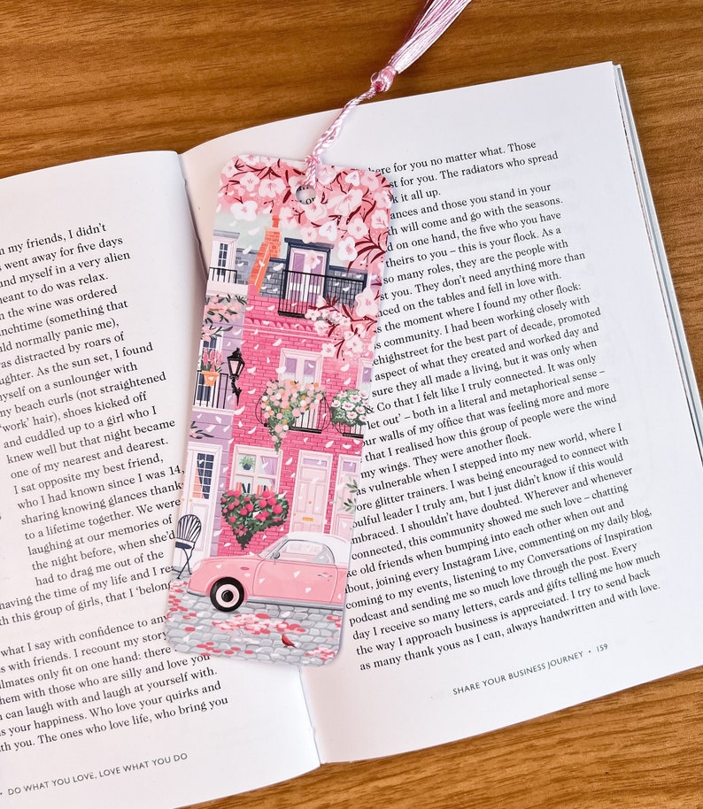 Pode incluir: Marcador de p&aacute;gina rosa e branco com um carro rosa e um design floral. O marcador mostra um carro rosa dirigindo por uma rua com edif&iacute;cios rosa e flores. O texto no marcador diz "Fa&ccedil;a o que ama, ame o que faz".