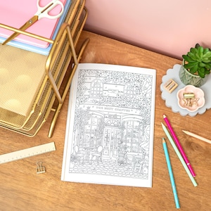 May include: An open coloring book with a detailed black and white illustration of a shop front, titled "The Next Chapter." Gold desk organizer, pink and blue pencils, and a succulent plant are also visible.