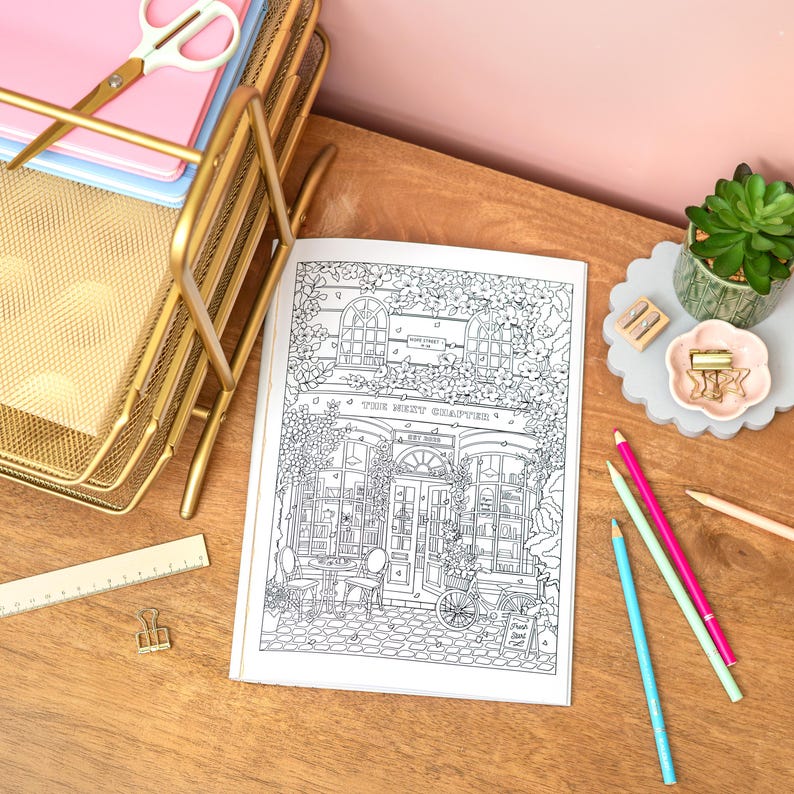May include: An open coloring book with a detailed black and white illustration of a shop front, titled "The Next Chapter." Gold desk organizer, pink and blue pencils, and a succulent plant are also visible.