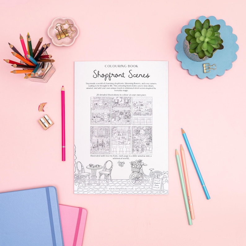 May include: A coloring book titled "Shopfront Scenes" with detailed illustrations of shop interiors and street scenes. The image includes colored pencils, a succulent plant, and stationery on a pink surface.