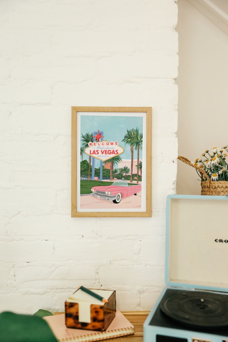 May include: A framed illustration of a pink car driving past the "Welcome to Fabulous Las Vegas, Nevada" sign with palm trees in the background.