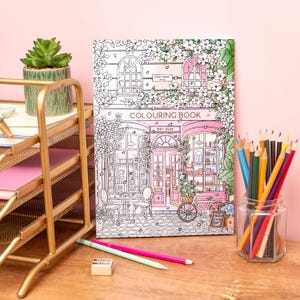 May include: A coloring book with a detailed illustration of a shopfront, featuring the text "COLOURING BOOK" and "EST 2020". The scene is in black and white with pink accents, surrounded by flowers. A jar of colored pencils and a gold desk organizer are also visible.