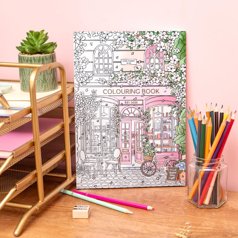 May include: A coloring book with a detailed illustration of a shopfront, featuring the text "COLOURING BOOK" and "EST 2020". The scene is in black and white with pink accents, surrounded by flowers. A jar of colored pencils and a gold desk organizer are also visible.