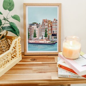 May include: A framed illustration of a canal scene in Amsterdam, Netherlands. The illustration features a canal with a boat and several buildings along the canal. The buildings are painted in shades of brown, red, and yellow. The sky is blue and there are trees in front of the buildings.