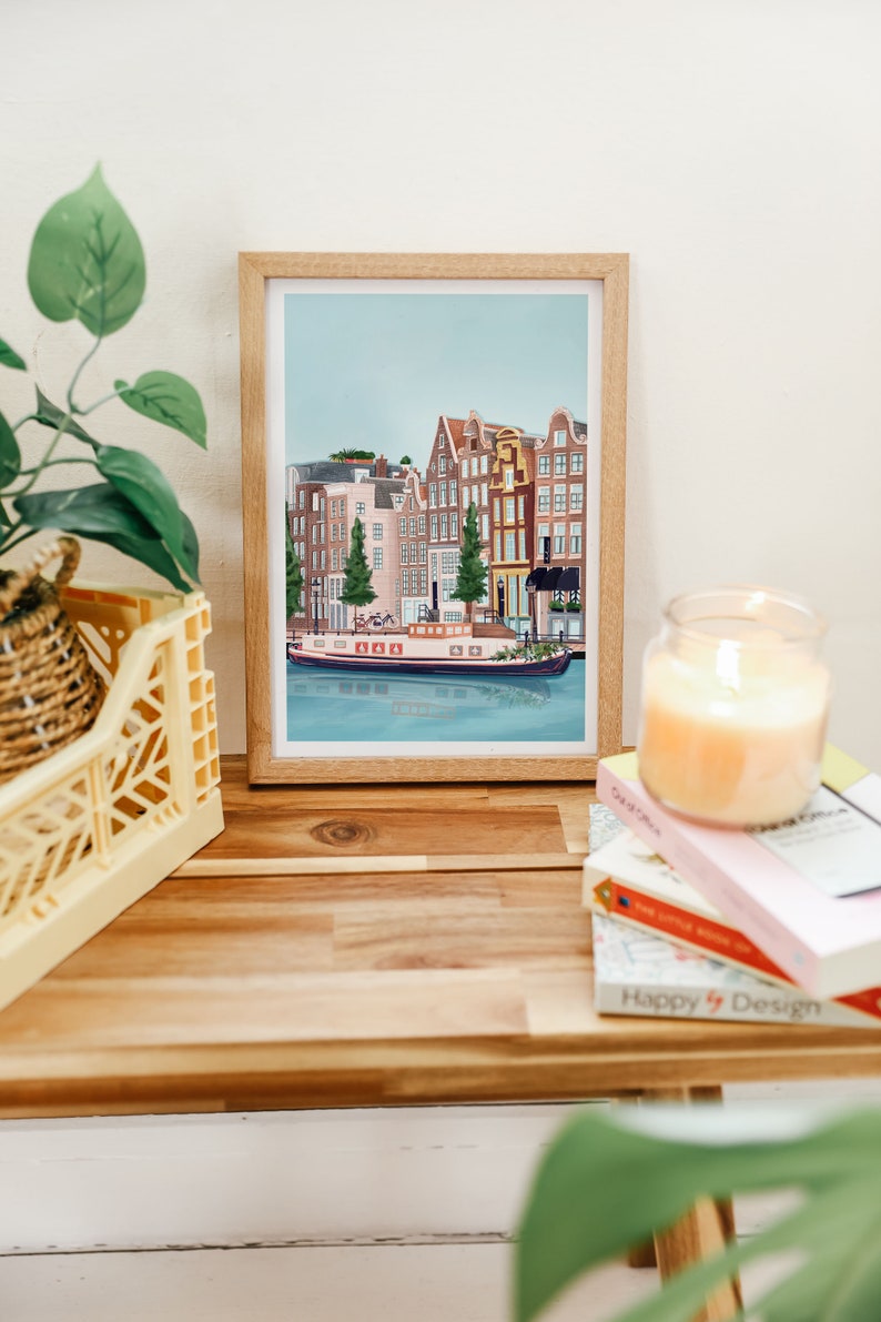 May include: A framed illustration of a canal scene in Amsterdam, Netherlands. The illustration features a canal with a boat and several buildings along the canal. The buildings are painted in shades of brown, red, and yellow. The sky is blue and there are trees in front of the buildings.