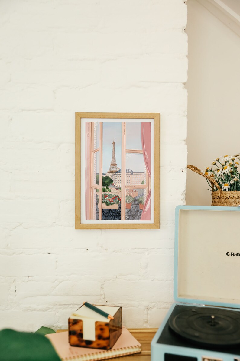 Op de afbeelding: Een ingelijst afdruk van een raamzicht met een roze gordijn en de Eiffeltoren in de verte. De afdruk bevindt zich op een witte bakstenen muur.