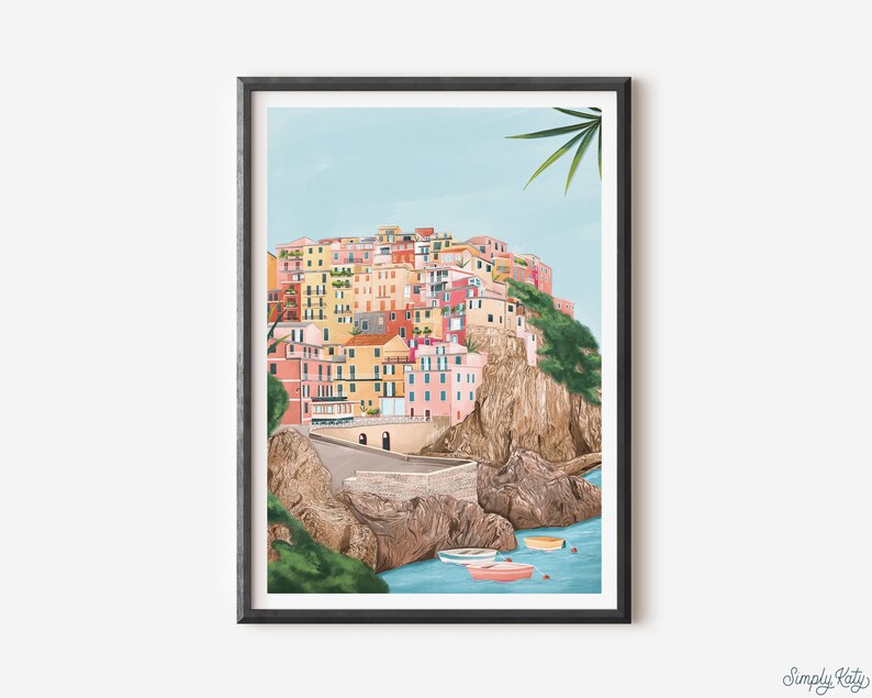 May include: A framed art print depicting a colorful coastal village nestled on a cliffside. The buildings are painted in shades of pink, yellow, and orange, with a light blue sky and turquoise water. Several small boats float in the water below.