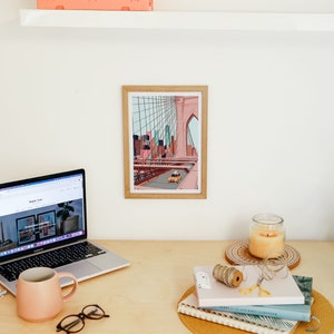 May include: A home office setup featuring a laptop, a framed art print of a bridge, a pink mug, and a candle. A small orange storage crate sits on a shelf above the desk. The desk is made of light-colored wood.
