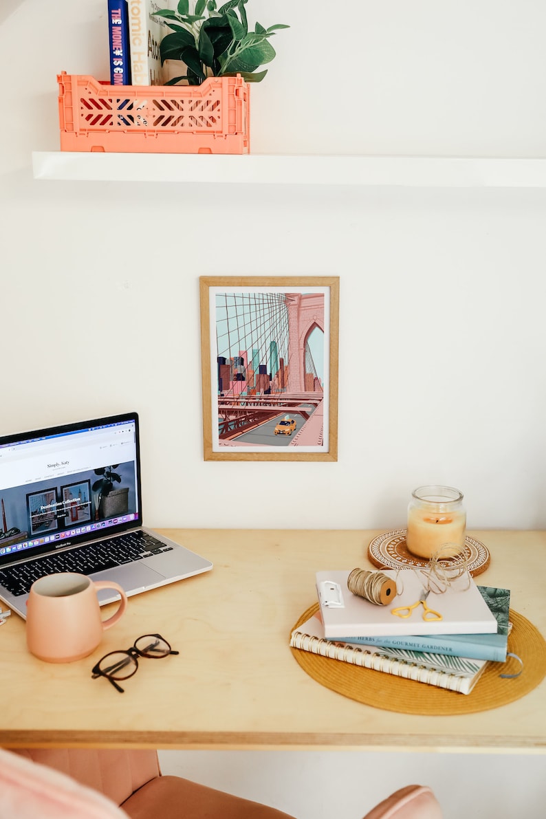 May include: A home office setup featuring a laptop, a framed art print of a bridge, a pink mug, and a candle. A small orange storage crate sits on a shelf above the desk. The desk is made of light-colored wood.