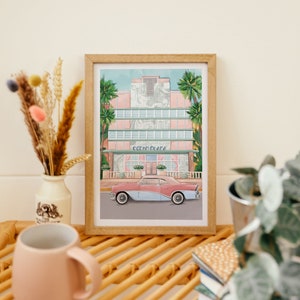 May include: A framed print of a pink and blue vintage car parked in front of a pastel pink building with palm trees. The building has the words "Ocean Plaza" on the front.