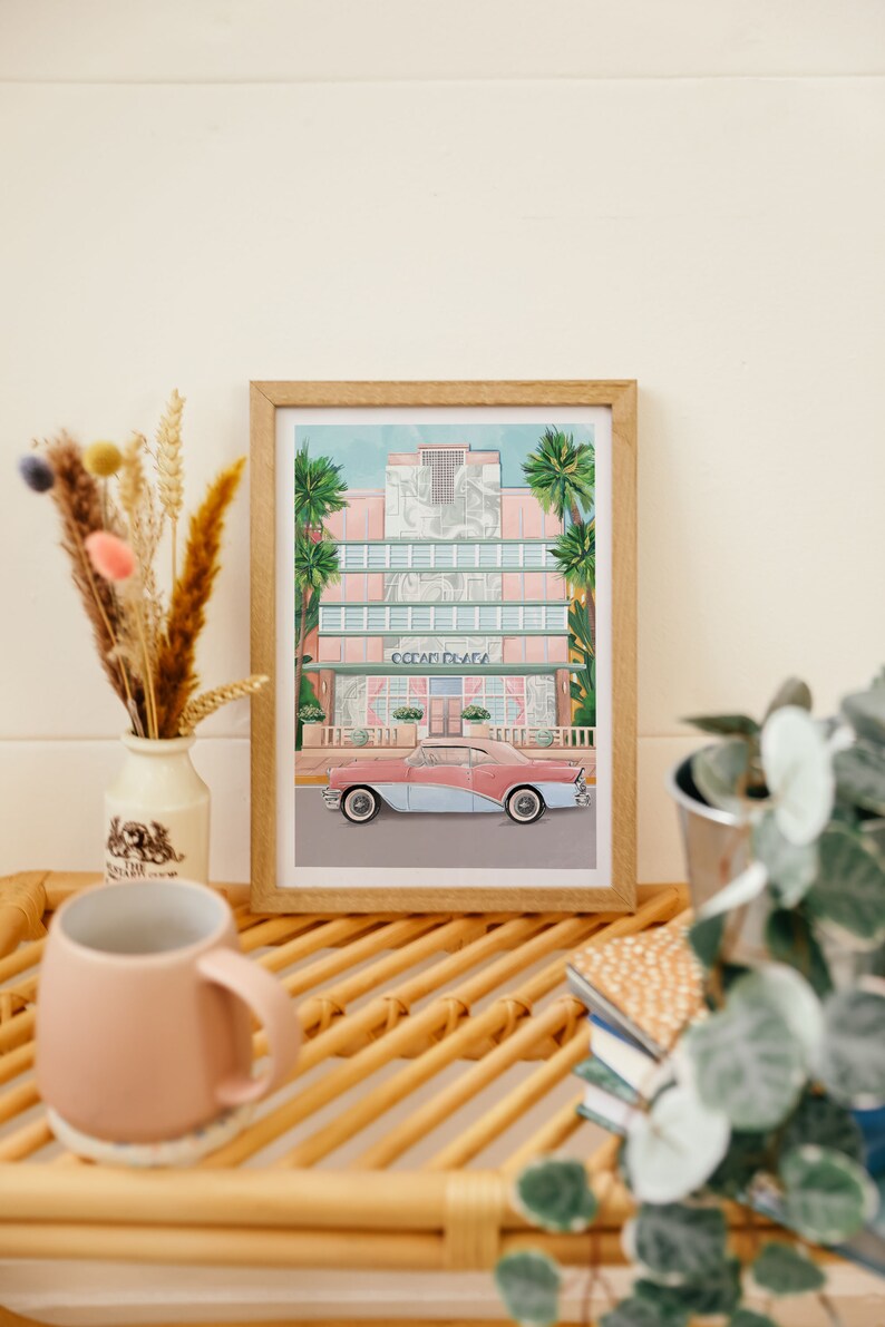 May include: A framed print of a pink and blue vintage car parked in front of a pastel pink building with palm trees. The building has the words "Ocean Plaza" on the front.