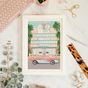 May include: A framed illustration of a pink and white building with the text "Ocean Plaza" above a vintage blue and pink car parked in front. The illustration is set against a blue sky with palm trees.