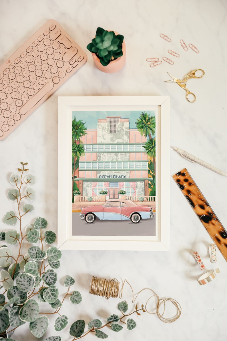May include: A framed illustration of a pink and white building with the text "Ocean Plaza" above a vintage blue and pink car parked in front. The illustration is set against a blue sky with palm trees.
