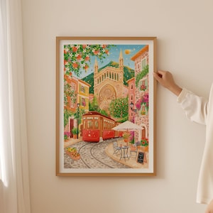 May include: A framed watercolor print depicts a European street scene with a red trolley car on cobblestone tracks. Buildings in shades of orange, yellow, and pink line the street, with a large church in the background. A cafe with a white umbrella and small table is visible.