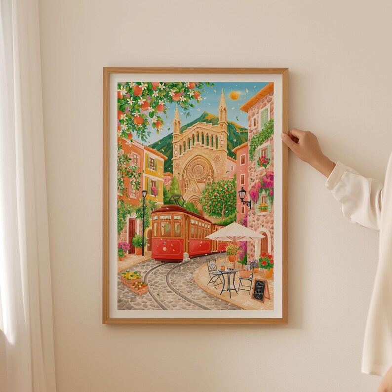 May include: A framed watercolor print depicts a European street scene with a red trolley car on cobblestone tracks. Buildings in shades of orange, yellow, and pink line the street, with a large church in the background. A cafe with a white umbrella and small table is visible.