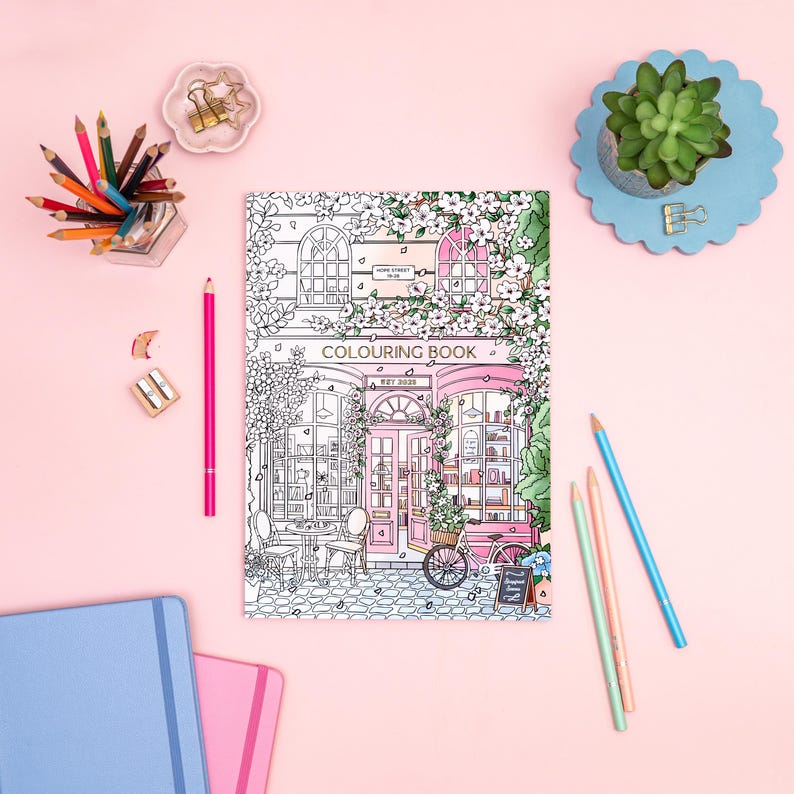 May include: A coloring book with a pink cover featuring a detailed line drawing of a shop front, with the text "COLOURING BOOK" and "EST 2023". The image is surrounded by colored pencils, a succulent, and notebooks on a pink background.