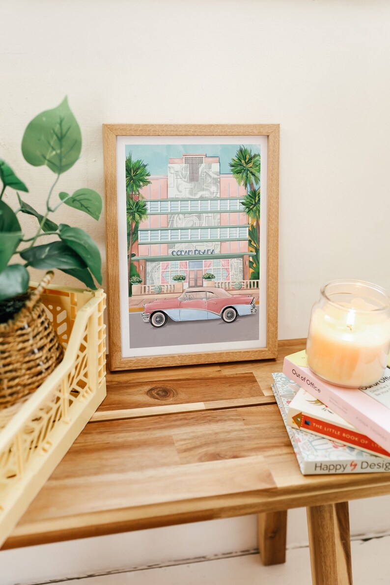 May include: A framed illustration of a pink and white building with the text "Ocean Plaza" above the entrance. A light blue and pink car is parked in front of the building. Palm trees are visible in the background.