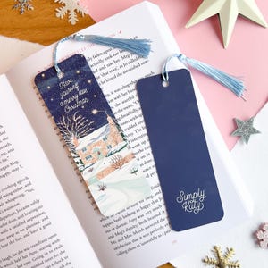 Op de afbeelding: Twee kerstboekmarkeringen met blauwe kwastjes liggen op een open boek. Eén bookmark toont een winterlandschap met een huis en de tekst "Have yourself a merry little Christmas". De andere is marineblauw met "Simply Katy" in wit.