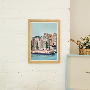 May include: A framed illustration of a canal scene in Amsterdam, Netherlands. The illustration features colorful buildings with red tile roofs, a canal with a boat, and trees. The frame is made of wood and has a light brown finish.