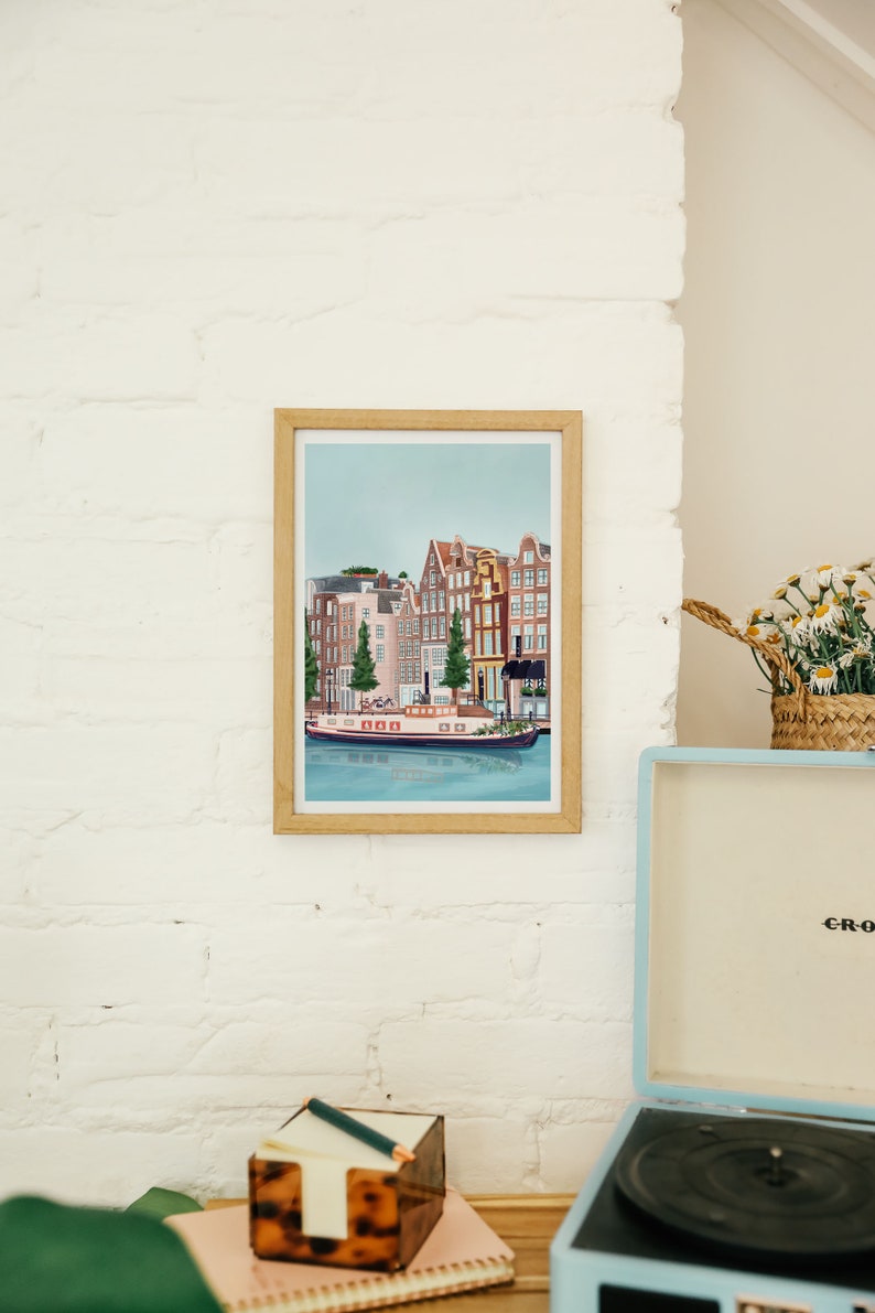 May include: A framed illustration of a canal scene in Amsterdam, Netherlands. The illustration features colorful buildings with red tile roofs, a canal with a boat, and trees. The frame is made of wood and has a light brown finish.