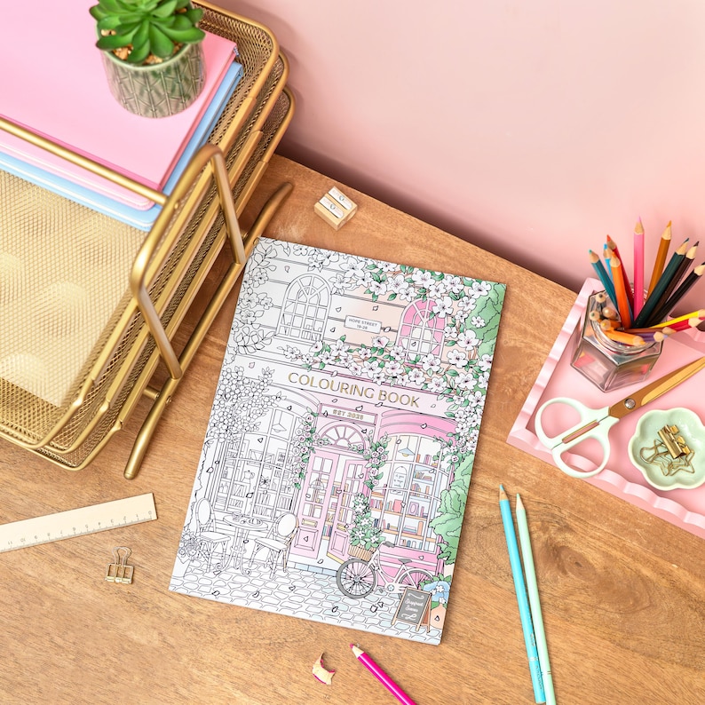 May include: A coloring book with a detailed line drawing of a shop front, featuring the text "COLOURING BOOK". The cover art is in black and white, with a pink door and floral accents. The book is on a wooden surface with stationery.