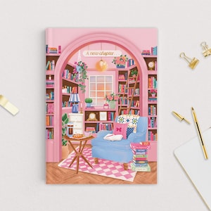 May include: A pink notebook with an illustration of a cozy library scene. The image features a blue armchair, bookshelves, and the text "A new chapter...". Gold pens and paper clips are on the white surface.