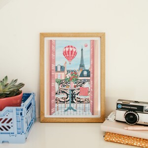 May include: A framed print of a Parisian balcony scene with a hot air balloon in the sky, a table with two chairs, and the Eiffel Tower in the background. The print has a pink and blue color scheme.