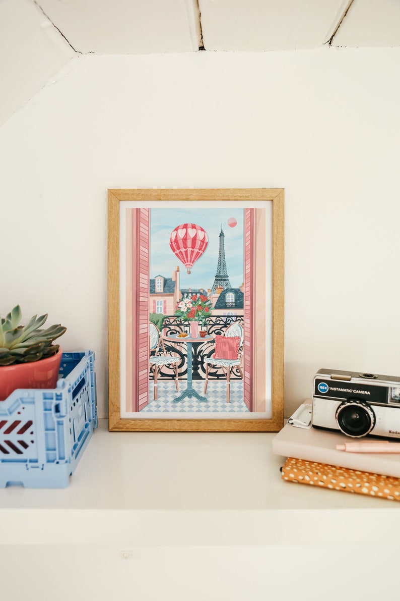May include: A framed print of a Parisian balcony scene with a hot air balloon in the sky, a table with two chairs, and the Eiffel Tower in the background. The print has a pink and blue color scheme.