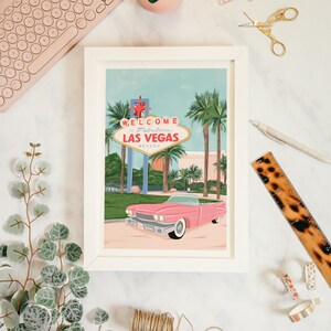 May include: A white framed print with a retro illustration of a pink convertible car driving past the iconic "Welcome to Fabulous Las Vegas" sign with palm trees in the background.
