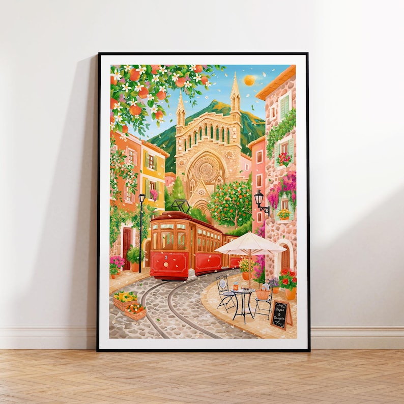 May include: A framed watercolor painting depicts a European street scene with a red trolley car on cobblestone tracks. Buildings in shades of orange, pink, and yellow line the street, with a large church in the background. A cafe with a white umbrella and small table is on the right.