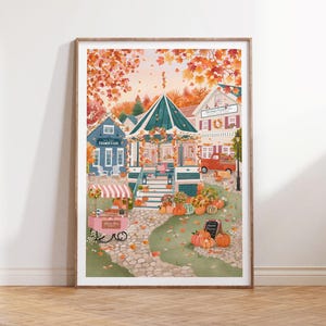 May include: A framed art print depicting an autumn town scene with a central gazebo, a blue creamery building, a red truck, and a pink market cart. The landscape is filled with orange and yellow fall foliage, pumpkins, and a stone path.
