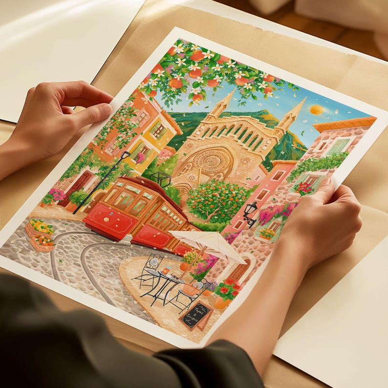 May include: A watercolor print depicting a European town scene with a red trolley car, cobblestone streets, and buildings in shades of orange, pink, and yellow. The artwork includes a church, cafe tables, and lush greenery, creating a vibrant and detailed illustration.