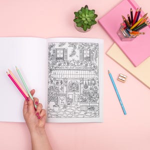 May include: An open coloring book with a detailed black and white illustration of a shop front, held by a person's hand. Pink, red, and green colored pencils are held in the hand. Other colored pencils, a succulent, and notebooks are on a pink surface.