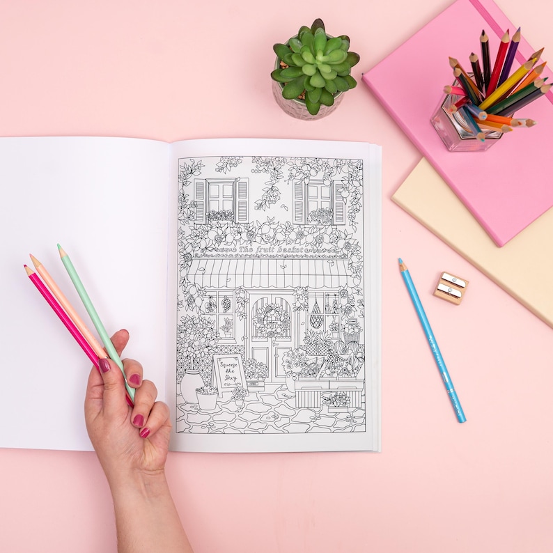 May include: An open coloring book with a detailed black and white illustration of a shop front, held by a person's hand. Pink, red, and green colored pencils are held in the hand. Other colored pencils, a succulent, and notebooks are on a pink surface.