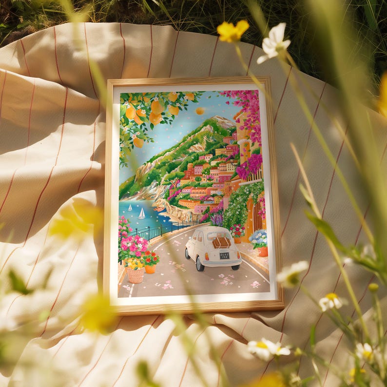 May include: A framed art print depicting a coastal Italian town with colorful buildings on a hillside. A white vintage car drives along a road lined with flowers. The artwork features lemons, sailboats, and vibrant pink and purple flowers.