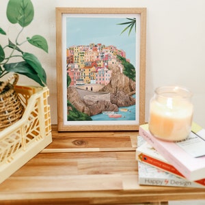 May include: A framed art print depicts a colorful coastal village scene with buildings in shades of pink, yellow, and orange. The print is in a light wood frame, resting on a wooden surface with a candle and books.
