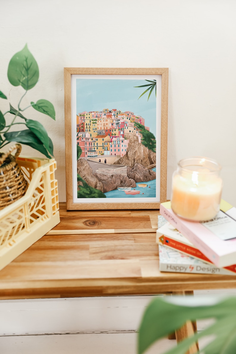 May include: A framed art print depicts a colorful coastal village scene with buildings in shades of pink, yellow, and orange. The print is in a light wood frame, resting on a wooden surface with a candle and books.
