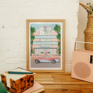 May include: A framed print of a pink art deco building with palm trees and a vintage car in front. The building has the words "Ocean Drive" on the front.