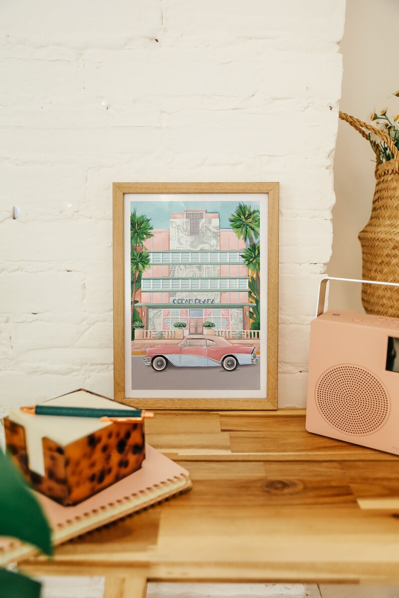 May include: A framed print of a pink art deco building with palm trees and a vintage car in front. The building has the words "Ocean Drive" on the front.