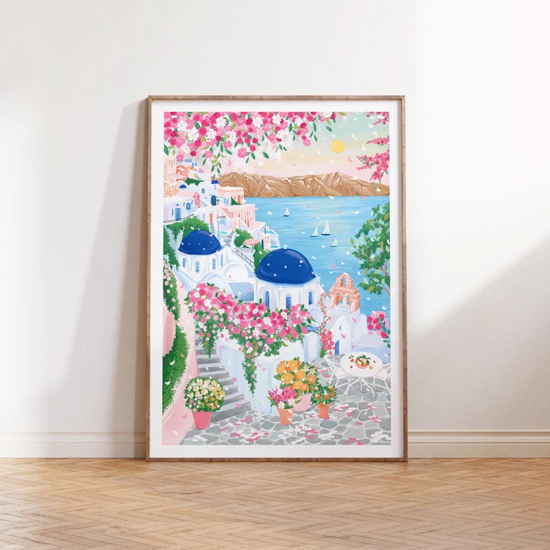 以下が含まれることがあります： 海岸沿いの村の風景を描いた額入りのアートプリント。 青いドームのある白い建物、鮮やかなピンクの花、ヨットのある海の景色が特徴です。 プリントは、薄い青い空と茶色の木製フレームを背景にしています。