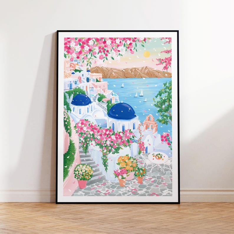 以下が含まれることがあります： 白い建物、青いドーム、ピンクの花が特徴の、絵のように美しい海岸沿いの村を描いた額入りのアートプリント。このアートワークは、穏やかな海、ヨット、遠くの山脈のある晴れた日を示しています。プリントは黒い縁で額装されています。
