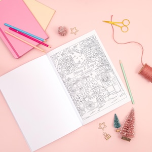 May include: An open coloring book with a detailed black and white illustration of a holiday scene, featuring a shop with the text "Merry & Bright Gift Boutique." Colored pencils, notebooks, and decorative items like miniature trees and stars surround the book on a pink surface.