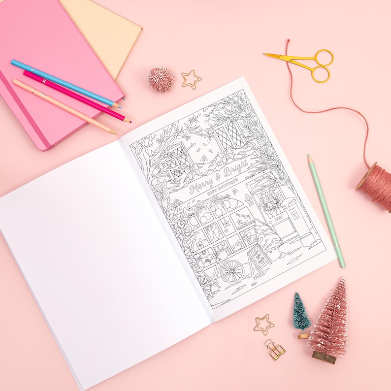 May include: An open coloring book with a detailed black and white illustration of a holiday scene, featuring a shop with the text "Merry & Bright Gift Boutique." Colored pencils, notebooks, and decorative items like miniature trees and stars surround the book on a pink surface.